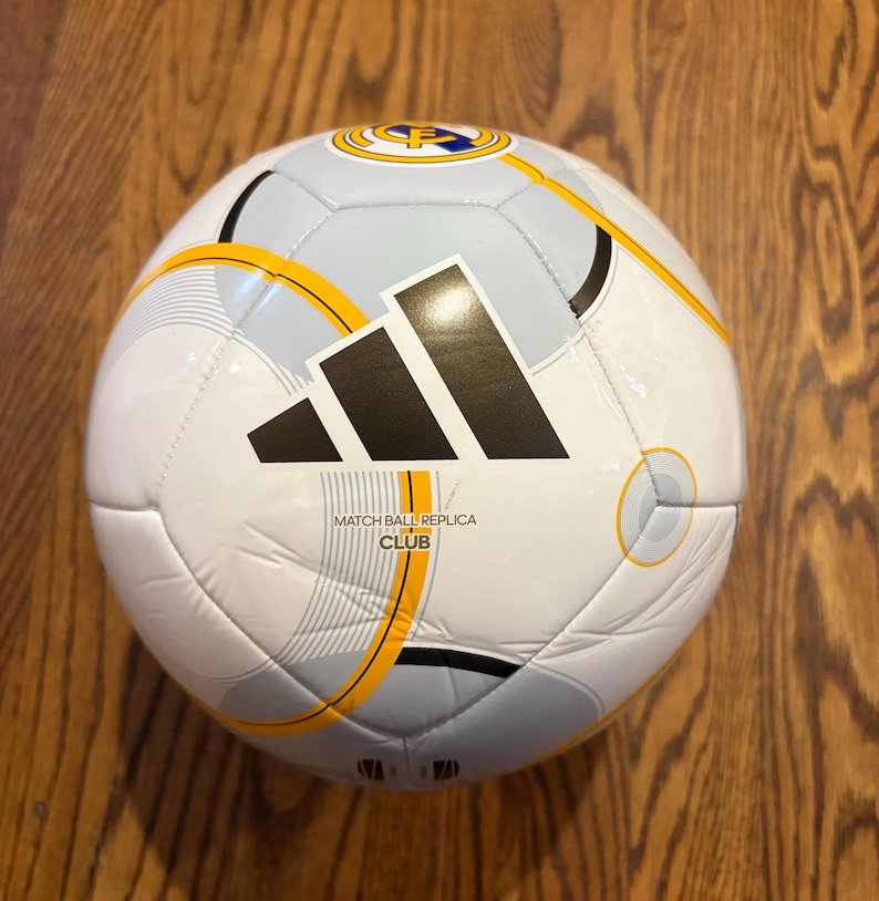 Puede incluir: Bal&oacute;n de f&uacute;tbol blanco y gris con el logo negro de Adidas y detalles amarillos. El bal&oacute;n tiene el texto "MATCH BALL REPLICA CLUB". Un logotipo del equipo es visible en la parte superior.
