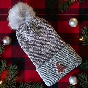 May include: A gray knit beanie with a faux fur pom-pom and a red and black plaid Christmas tree patch. The hat is set against a red and black plaid background with silver ornaments and pine branches.