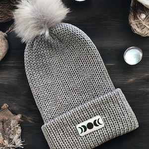 May include: Gray knitted beanie with a faux fur pom pom and a silver patch with a moon phase design.