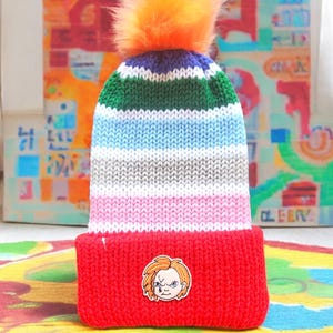 May include: A red knit beanie with a striped pattern in green, blue, grey, white, and pink. The beanie has a fluffy orange pom pom on top and a small embroidered image of a character with red hair and a mischievous expression.