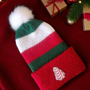 May include: A knitted winter hat with a white faux fur pom-pom. The hat features red, green, and white stripes, with a small Christmas tree patch on the red cuff. The hat is displayed on a red surface with Christmas decorations.