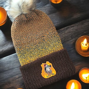 May include: A brown and yellow knit beanie with a fluffy pom pom on top. The beanie has a brown cuff with an embroidered patch of a dog in a fire saying "This is fine".