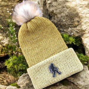 May include: A mustard yellow knit beanie with a light beige fold-over brim. The hat features a fluffy, light pink and gray pom-pom and a black Bigfoot patch. The hat is resting on a rocky surface.