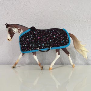 May include: A toy horse with a black blanket featuring a colorful star pattern and a bright blue trim. The horse has a brown and white coat, with a light-colored mane and tail.