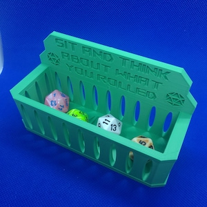 May include: A green plastic dice tray with the text "Sit and think about what you rolled" on the front. The tray has a grid of slots to hold dice. There are five dice in the tray, including a pink d6, a green d6, a blue d12, a white d10, and a brown d6.
