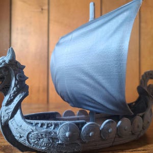 May include: A blue and grey 3D printed model of a Viking longship with a dragon head prow and a sail. The ship has a detailed design with shields and other decorative elements.