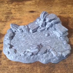 Puede incluir: Una pieza de terreno en miniatura de color gris, impresa en 3D, que representa un afloramiento rocoso con una pequeña área plana en el centro. La pieza está diseñada para su uso en juegos de mesa o juegos de guerra en miniatura.