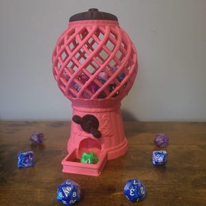 May include: A pink gumball machine with a brown lid and a clear, wire-mesh sphere filled with colorful dice. The machine has a crank on the side and a slot at the bottom for dispensing dice. There are several dice scattered on a wooden surface.