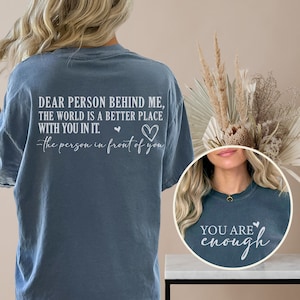 May include: A blue t-shirt with the text "Dear person behind me, the world is a better place with you in it. -the person in front of you" and two hearts on the back. The front of the shirt has the text "You are enough" and a heart.