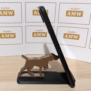 Pode incluir: Um smartphone preto repousa sobre um suporte de telefone em forma de cão preto e castanho. O cão é castanho e parece ser da raça pointer. O suporte é feito de madeira ou material semelhante. O fundo apresenta um padrão repetido com o texto "maker AMW".