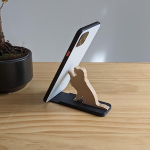 May include: A white smartphone propped up in a black and wood phone stand shaped like a dog. The stand is on a light wood surface.