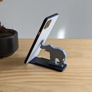May include: A white smartphone propped up on a black stand with a grey elephant figurine. The elephant is holding its trunk up in the air.