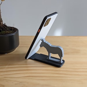 Puede incluir: Un teléfono inteligente blanco está apoyado en un soporte de teléfono negro con forma de burro. El soporte está sobre una superficie de madera marrón claro.