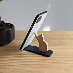 May include: A white smartphone is propped up in a black phone stand with a wooden cat figurine reaching up towards the phone.