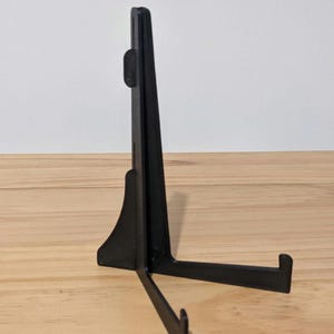 Puede incluir: Un soporte de exhibición negro que cuenta con un respaldo vertical con una pequeña pestaña ajustable y dos brazos horizontales que se extienden hacia adelante para sostener objetos. La estructura es de material mate y sirve de apoyo.