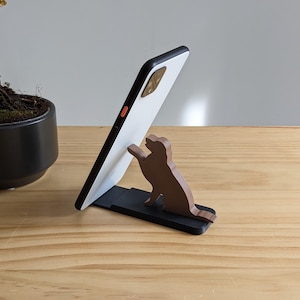May include: A white smartphone propped up on a wooden phone stand shaped like a dog. The stand is on a light brown wooden surface.