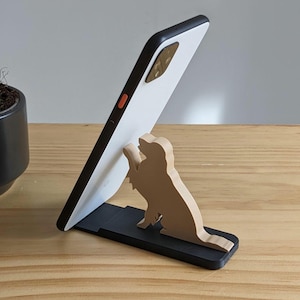 May include: A black smartphone stand featuring a light tan silhouette of a dog sitting with one paw raised. The stand is constructed with a flat rectangular base and a vertical dog-shaped support piece that holds a white smartphone at an angle.