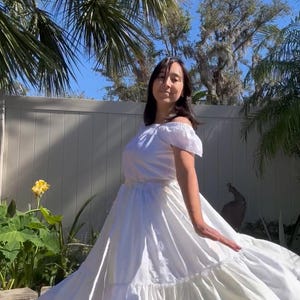 K&ouml;nnte beinhalten: Eine Frau in einem wei&szlig;en, schulterfreien Kleid mit einem ger&uuml;schten Rock und einer Spitzenborte. Sie steht in einem Garten mit Palmen und einem wei&szlig;en Zaun im Hintergrund.