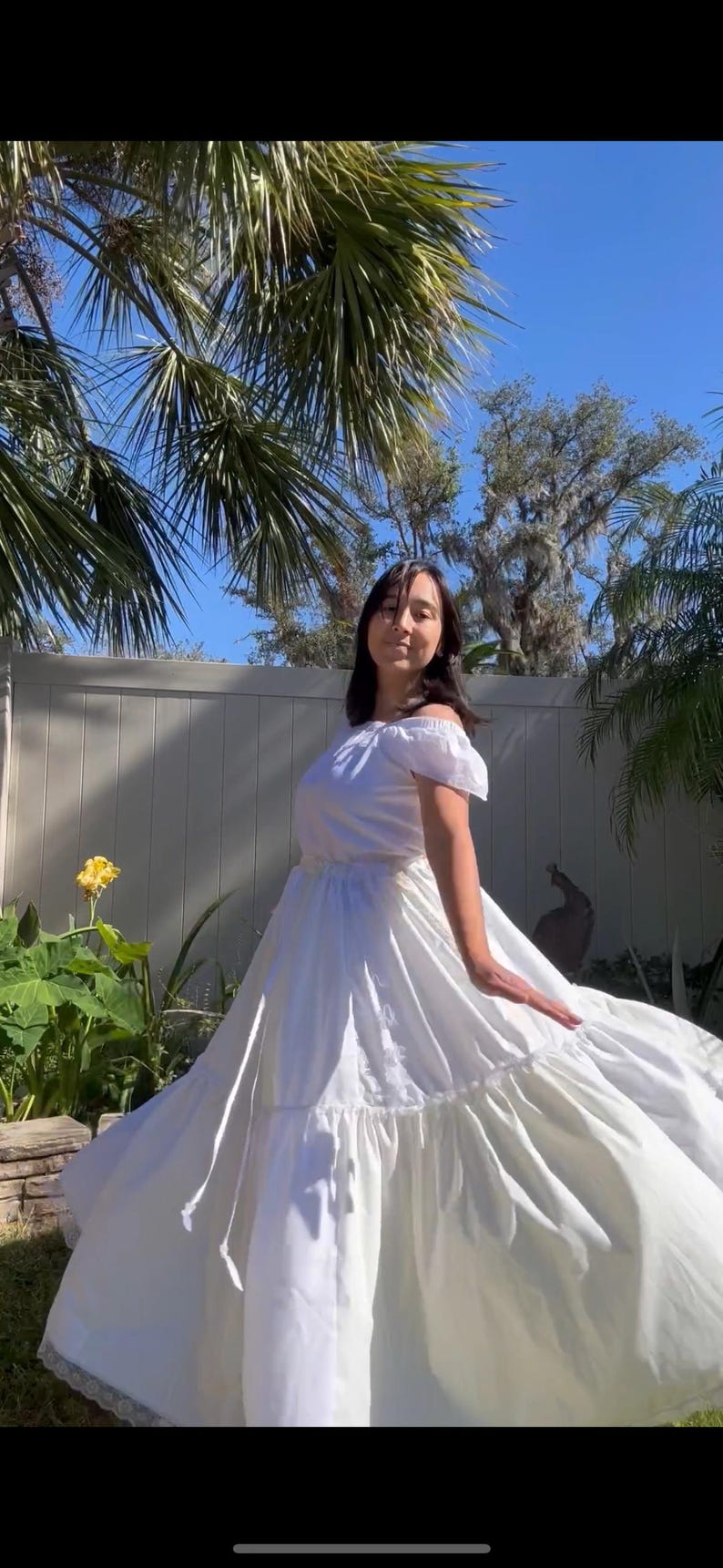 K&ouml;nnte beinhalten: Eine Frau in einem wei&szlig;en, schulterfreien Kleid mit einem ger&uuml;schten Rock und einer Spitzenborte. Sie steht in einem Garten mit Palmen und einem wei&szlig;en Zaun im Hintergrund.