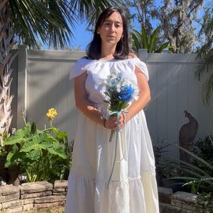 K&ouml;nnte beinhalten: Eine Frau in einem wei&szlig;en Kleid mit Spitzenbesatz h&auml;lt einen Strau&szlig; aus blauen und wei&szlig;en Blumen. Sie steht vor einem wei&szlig;en Zaun mit gr&uuml;nem Gr&uuml;n hinter ihr.