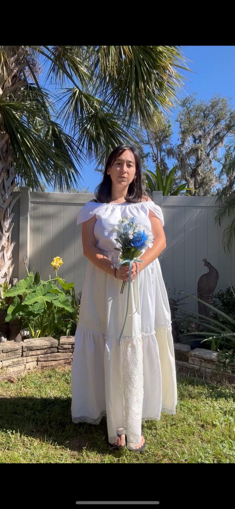 K&ouml;nnte beinhalten: Eine Frau in einem wei&szlig;en Kleid mit Spitzenbesatz h&auml;lt einen Strau&szlig; aus blauen und wei&szlig;en Blumen. Sie steht vor einem wei&szlig;en Zaun mit gr&uuml;nem Gr&uuml;n hinter ihr.