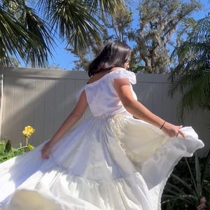 K&ouml;nnte beinhalten: Ein wei&szlig;es Kleid mit einem ger&uuml;schten Rock und einer Spitzenborte. Das Kleid wird von einer Person in einem Garten getragen.