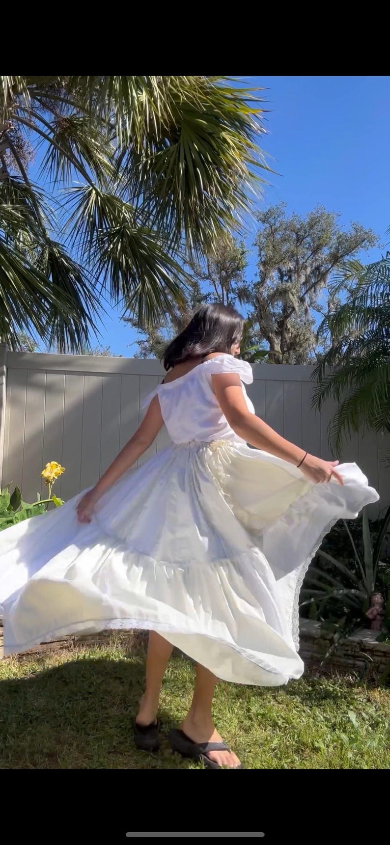 K&ouml;nnte beinhalten: Ein wei&szlig;es Kleid mit einem ger&uuml;schten Rock und einer Spitzenborte. Das Kleid wird von einer Person in einem Garten getragen.
