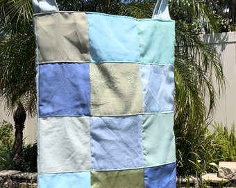 May include: A patchwork tote bag constructed from square fabric panels in various shades of blue, light green, and sage. The bag features a long, light blue shoulder strap and a rectangular body with a grid-like pattern of stitched fabric squares.