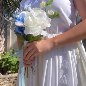 K&ouml;nnte beinhalten: Ein wei&szlig;es, schulterfreies Kleid mit einer Spitzenborte und einem blauen Band, das um einen Strau&szlig; wei&szlig;er und blauer Blumen gebunden ist.