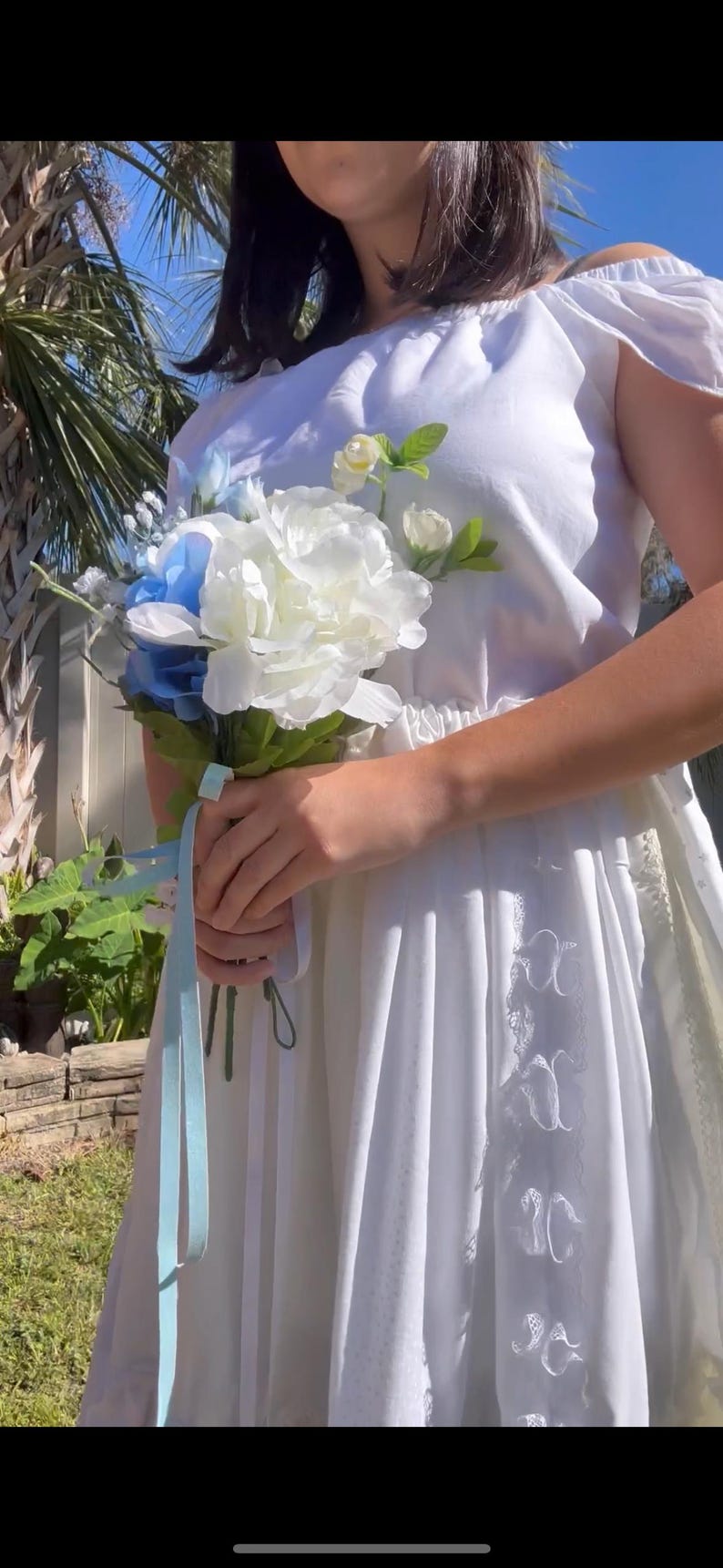 K&ouml;nnte beinhalten: Ein wei&szlig;es, schulterfreies Kleid mit einer Spitzenborte und einem blauen Band, das um einen Strau&szlig; wei&szlig;er und blauer Blumen gebunden ist.