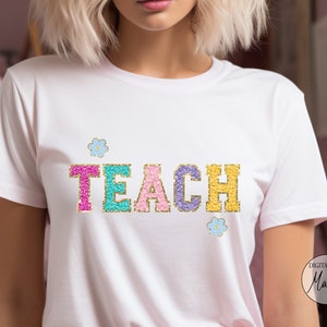 May include: White t-shirt featuring the word "TEACH" in colourful, glittery letters. The letters are outlined in gold and decorated with small flower graphics. A casual style t-shirt, suitable for teachers or educators.