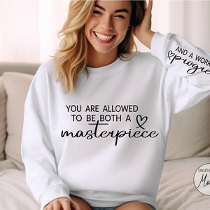 May include: White sweatshirt with the text "YOU ARE ALLOWED TO BE BOTH A masterpiece" and a heart symbol. The right sleeve has the text "AND A WORK IN progress" with a heart symbol.
