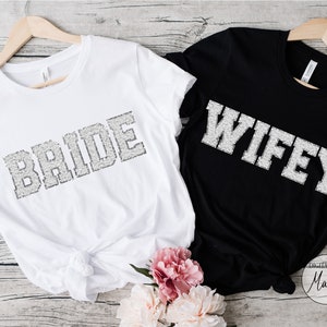 Peut inclure: Deux t-shirts blancs avec les mots "BRIDE" et "WIFEY" imprimés en paillettes argentées sur fond noir. Les t-shirts sont suspendus à des cintres en bois et sont affichés sur une surface en bois.