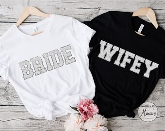 May include: Two white t-shirts with the words "BRIDE" and "WIFEY" printed in silver glitter on a black background. The shirts are hanging on wooden hangers and are displayed on a wooden surface.