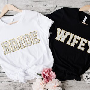 May include: Two white t-shirts with gold glitter lettering. One shirt says "BRIDE" and the other says "WIFEY".