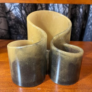 May include: A decorative, three-sectioned container with a unique, flowing design. The container is made of a speckled, two-toned material, with a light tan color at the top that fades into a darker brown at the base. The design features two cylindrical cups and a central, curved section.