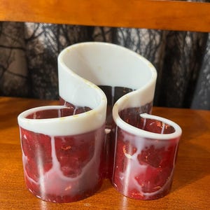 May include: A set of three decorative candle holders. The holders are red with white accents and a glossy finish. The design features a unique, curved shape, adding a modern touch to any space. The candle holders are made of a durable material.