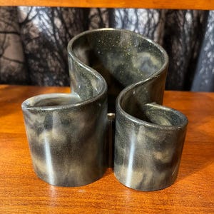 May include: A black and gold decorative pen holder with three compartments. The holder has a unique, curved design and sits on a wooden surface. The interior of the holder is black, and the exterior has a gold, speckled pattern.