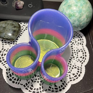May include: A colorful, three-section resin container with a blue exterior, transitioning to pink and green bands inside. The interior has a gold shimmer. The container sits on a white lace doily, with a green sphere and other stones in the background.
