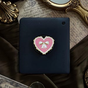 May include: A black e-reader with a pink heart-shaped PopSocket with a white bow and gold trim. The e-reader is on a dark brown surface with antique paper and a gold-framed mirror. The text "SIX WONDERS OF THE WORLD" is visible.