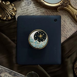May include: A navy blue Rakuten Kobo e-reader with a decorative pop socket featuring a celestial design. The pop socket has a gold border, a crescent moon, stars, and stylized clouds in shades of blue and white. The background includes vintage ephemera.