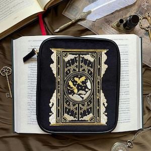 May include: A black zippered pouch with gold embroidery depicting a door, clouds, and a dragon. The pouch rests on an open book. A white feather quill and a pocket watch are also visible in the image.