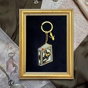 May include: A gold-framed keychain with a miniature book design. The book has a celestial scene on the cover. The keychain has a gold ring and a charm of a flying figure. The background is black.
