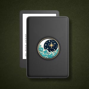 May include: A black e-reader device featuring a circular accessory on its back. The accessory displays a gold-rimmed design of a white crescent moon, a dark blue night sky with gold stars, and stylised teal and white clouds at the bottom.