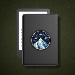 May include: A black e-reader device featuring a circular adhesive grip attached to its back. The grip displays a gold-rimmed illustration of blue mountain peaks set against a dark navy night sky filled with gold stars and sparkling constellations.