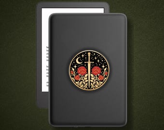 May include: A black e-reader device featuring a circular sticker on its back. The sticker displays a gold sword centered between red roses and gold leaves, set against a black background with a crescent moon and small stars in the upper section.