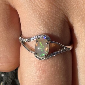 Könnte beinhalten: Ein silberner Ring mit einem weißen Opal-Edelstein, der von kleinen klaren Steinen umgeben ist.