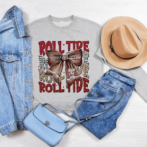 May include: A gray sweatshirt with the words "Roll Tide" printed in red and black. The sweatshirt features a red and white bow with a small white elephant charm. The sweatshirt is paired with a blue denim jacket, blue jeans, a light blue purse, and a brown fedora.
