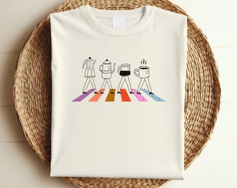 May include: White t-shirt with a graphic of coffee-themed characters walking across a rainbow-colored crosswalk. The characters include a coffee pot, a cup of coffee, and a mug.