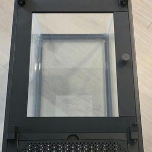 May include: Black terrarium with a clear glass viewing area. The terrarium has a rectangular shape with a hinged door and ventilation grilles. The bottom of the terrarium has the text "CEX".
